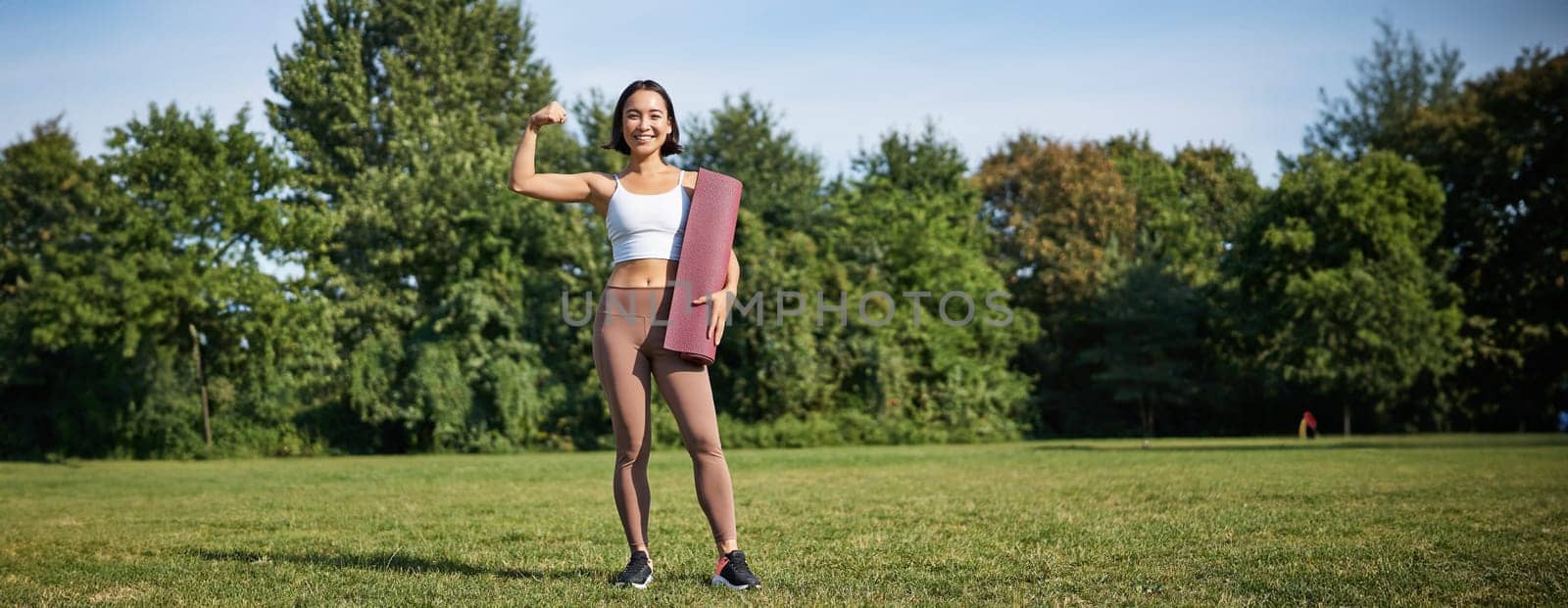 Confident and sporty young asian woman, standing in park with rubber mat, wearing sportswoman, smiling at camera, workout on fresh air.