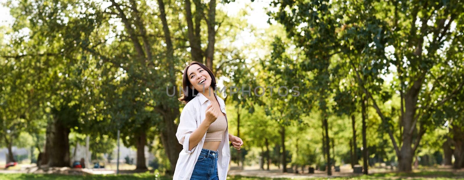 Happy young asian woman walking alone, dancing and singing in park, smiling carefree. People and lifestyle concept.