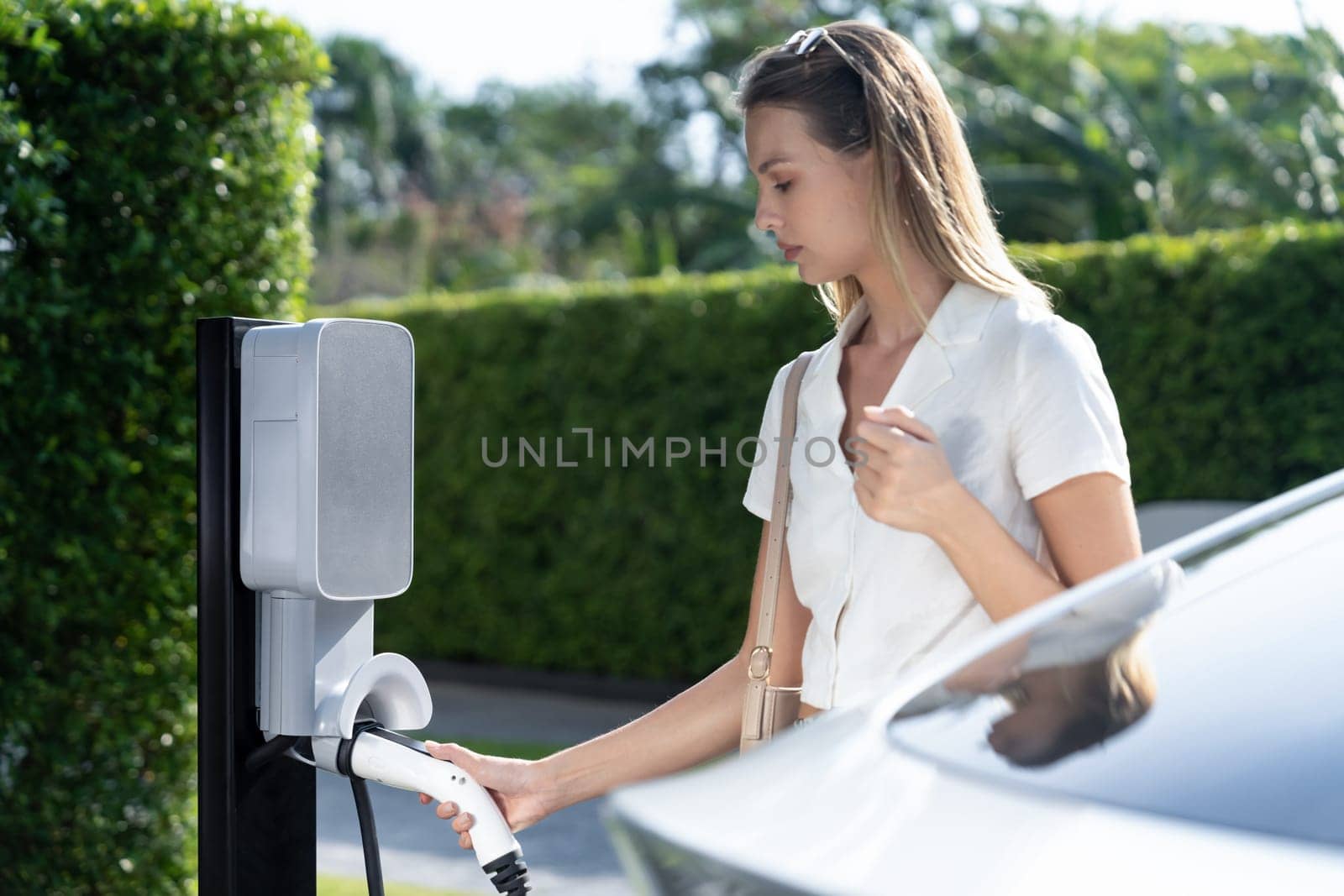 Young woman travel with EV electric car charging in green sustainable city outdoor garden in summer. Urban sustainability lifestyle by green clean rechargeable energy of electric BEV vehicle innards