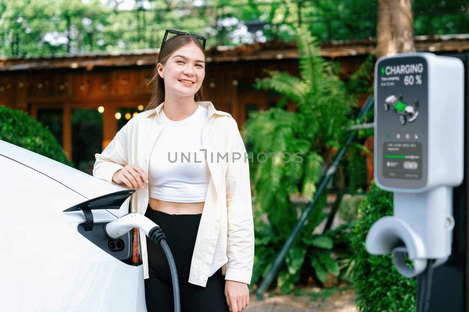 Sustainable urban commute with young woman pay electricity for EV electric car recharging at outdoor cafe in springtime garden, green city sustainability and environmental friendly EV car. Expedient