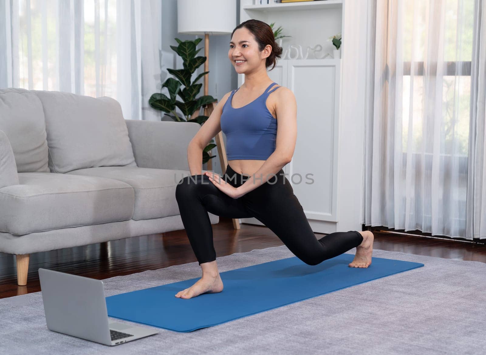 Asian woman in sportswear doing yoga exercise on fitness mat as her home workout training routine. Healthy body care lifestyle woman watching online yoga video on laptop. Vigorous