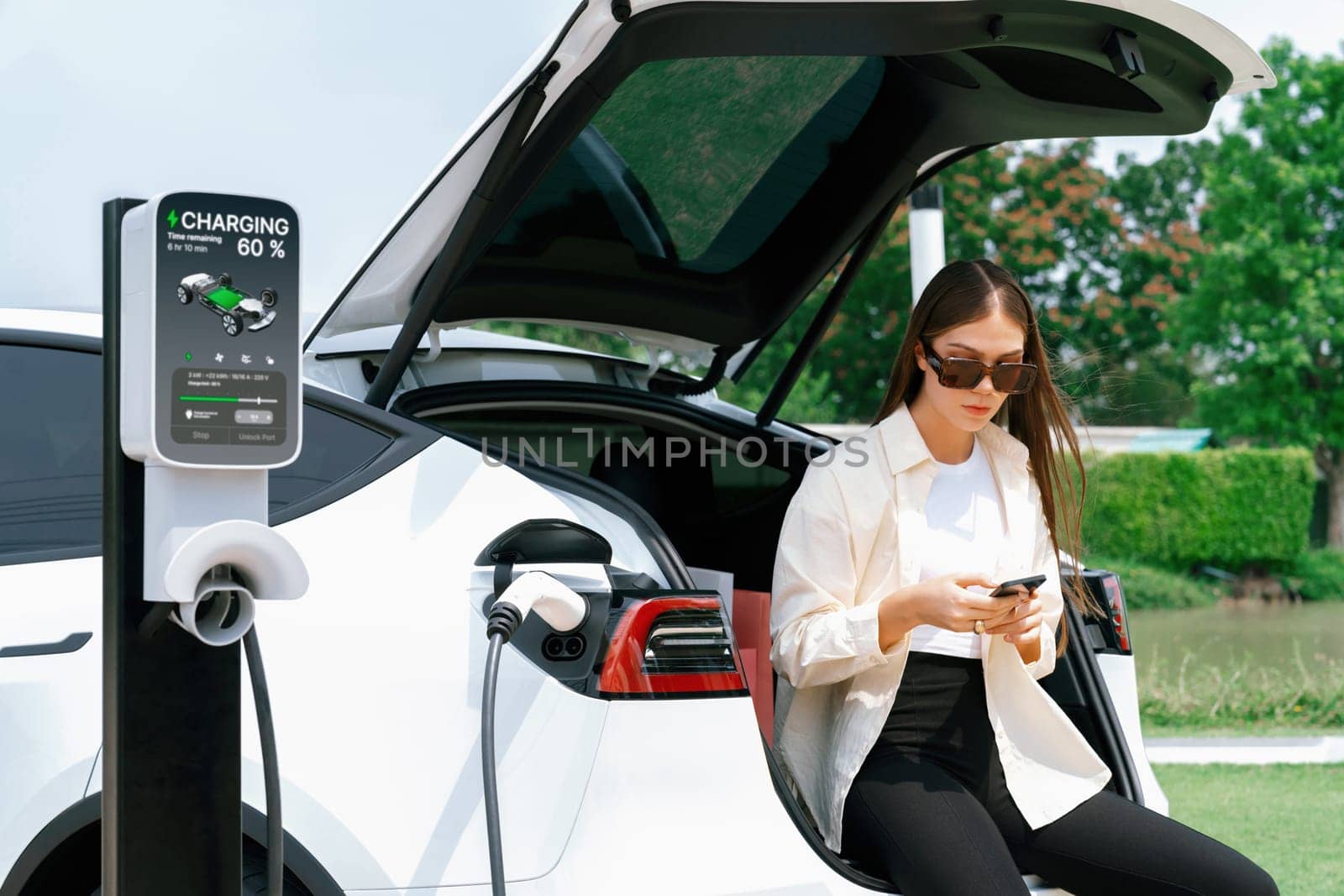 Young woman use smartphone to pay for electricity at public EV car charging station green city park. Modern environmental and sustainable urban lifestyle with EV vehicle. Expedient