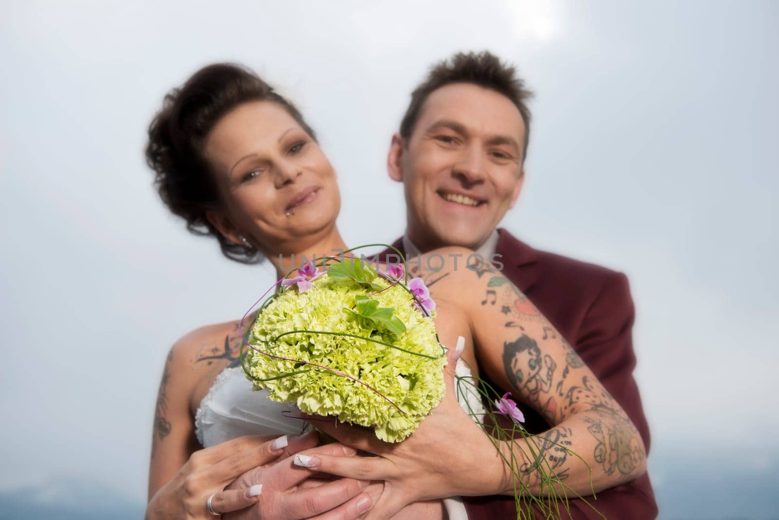 A bride with tattoos shows her bridal bouquet,behind it her groom