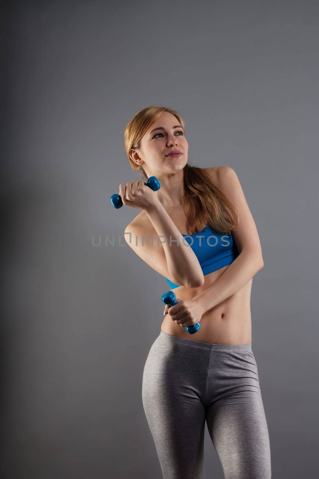 Young sportswoman with blue fitness dumbbells against gray background