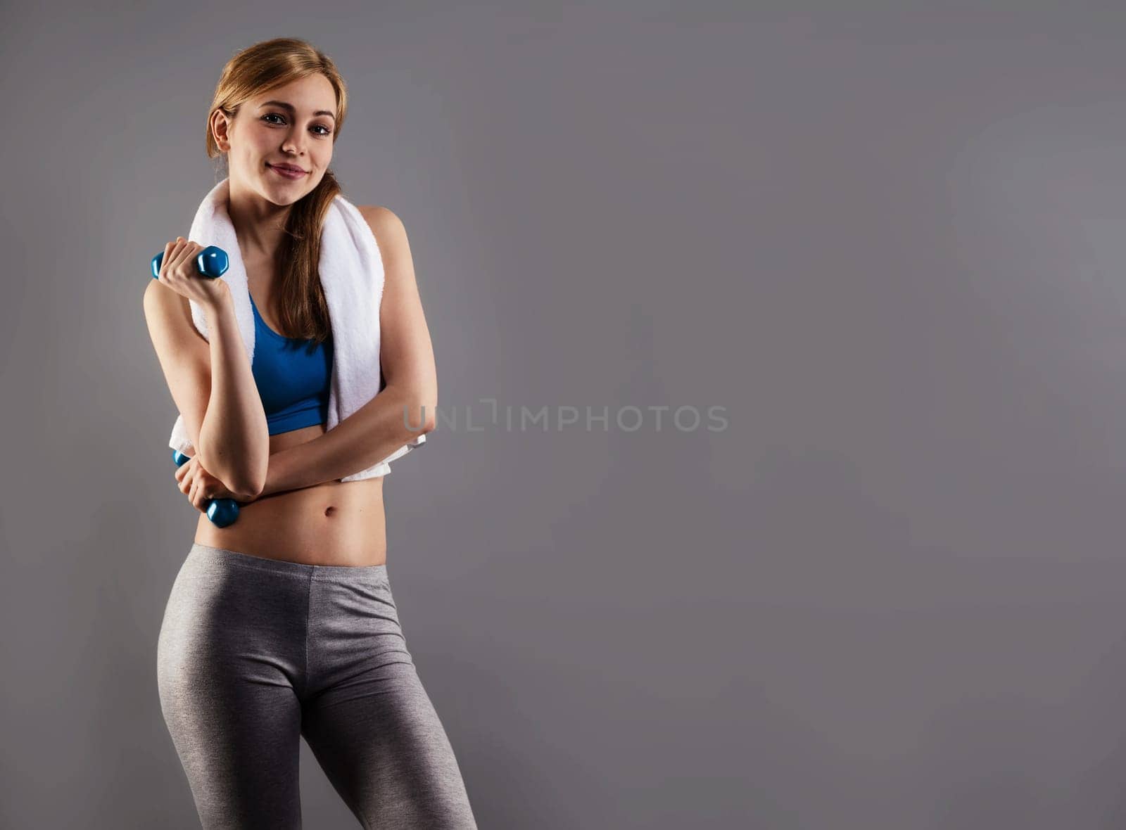 Young sportswoman with white towel and fitness dumbbells against gray background