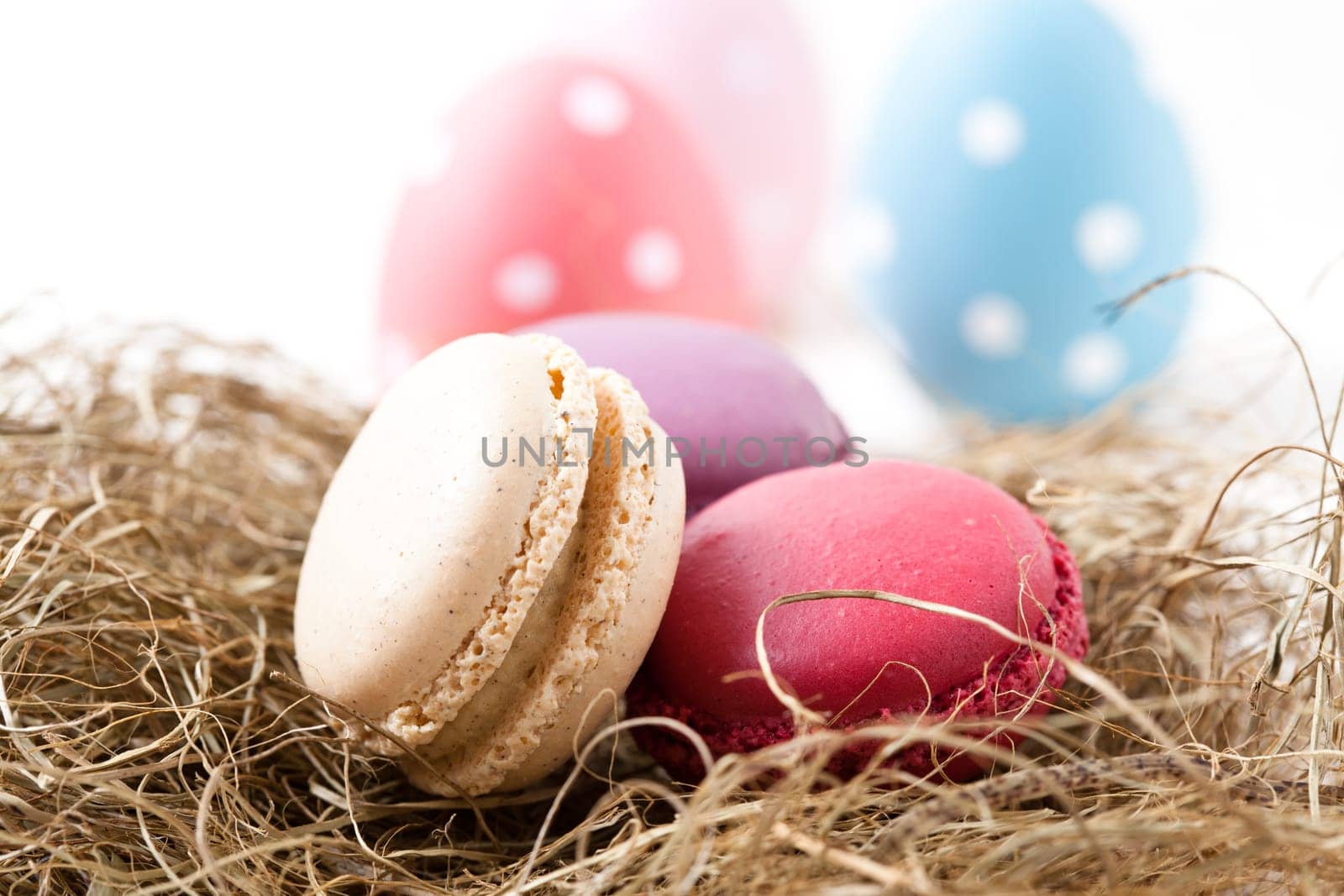 Close-up of colorful macaroons resting in straw nest and Easter eggs on light background