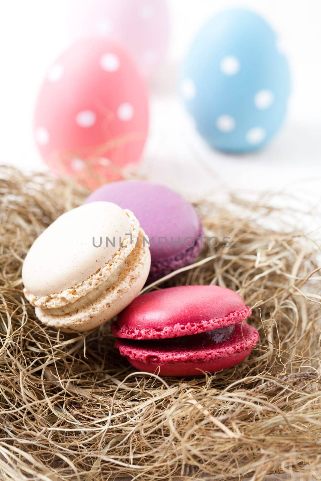 Close-up of colorful macaroons resting in straw nest and Easter eggs on light background