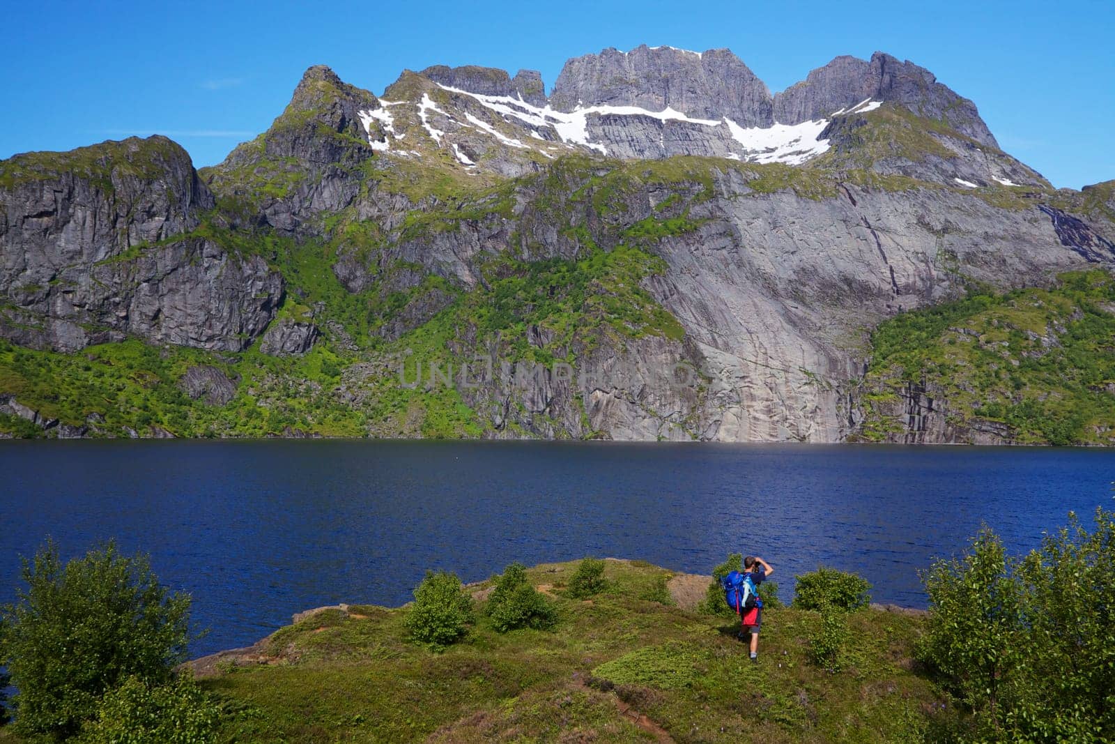 Young hiker with backpack standing by fjord on Lofoten islands in Norway on sunny day