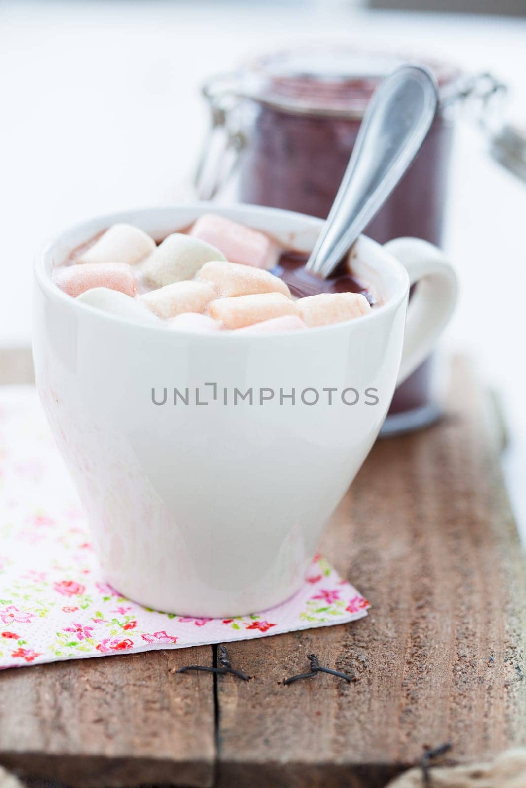 Closeup of cup of hot chocolate with coloured marshmallows resting on serviette on wooden table with glass jar of cocoa powder in background
