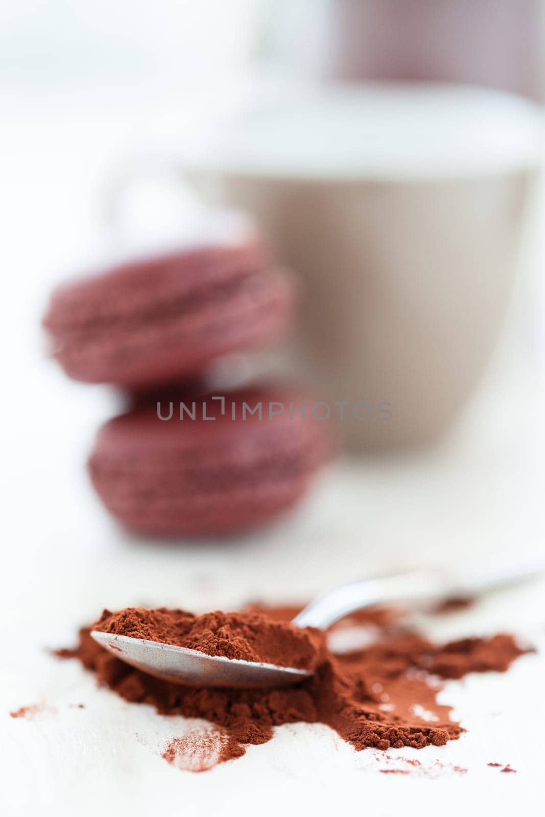 Closeup of cocoa powder and spoon with two macaroons and cup and glass jar in background