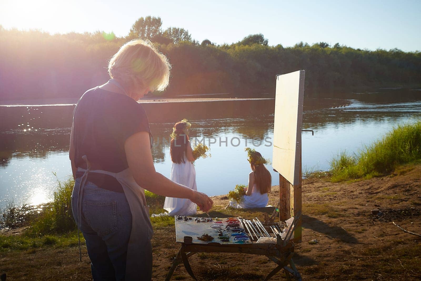 Adult female artist painting picture near water of river or lake in nature and girls in white sundress and flower wreath. Artist and models posing in holiday of Ivan Kupala in nature at sunset