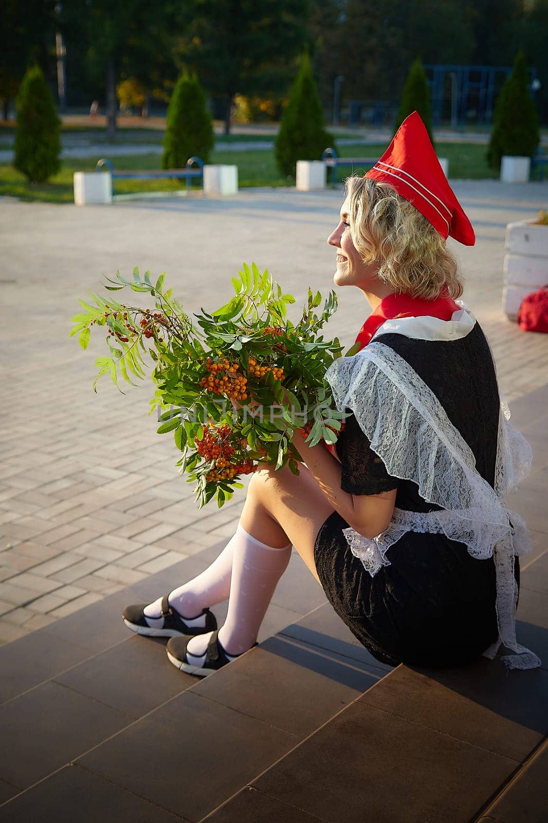 A Girl in black school uniform, white apron and red tie on steps of school with bouquet of flowers. Nostalgia photo shoot of teenager of female pioneer from USSR costume for September 1 or graduation