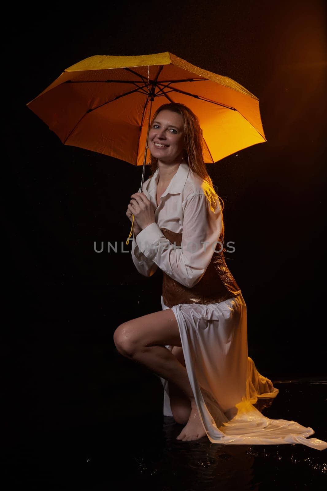 Beautiful sexy skinny girl in a dark hall with splashes of water and yellow umbrella under rain. Female Model blonde woman posing with water on black background in the studio