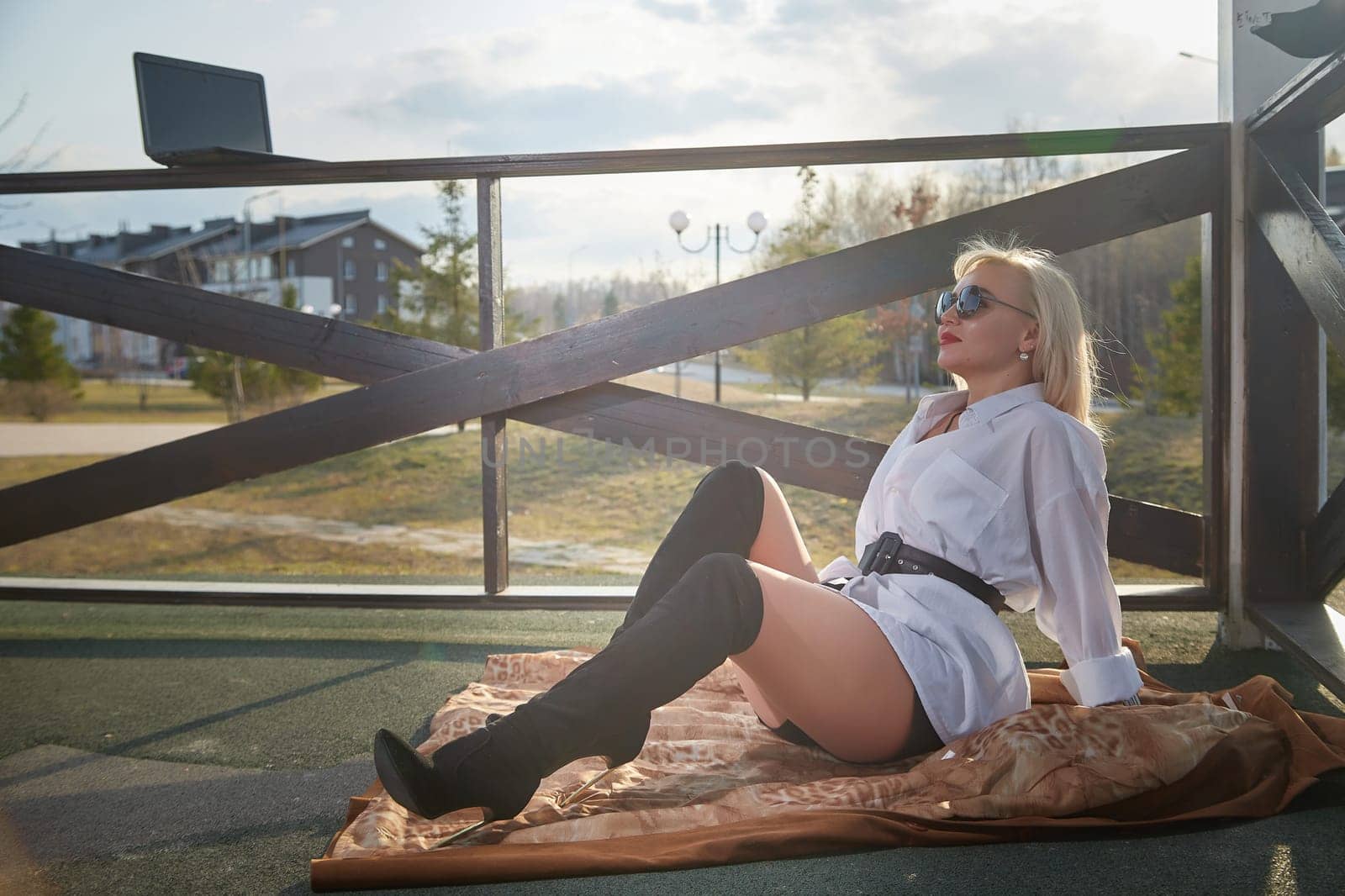 Fashionable beautiful blonde girl in a white shirt and a short skirt is resting in a gazebo with a laptop on sunny autumn, spring day. Lazy and relaxing businesswoman, manager, student in nature