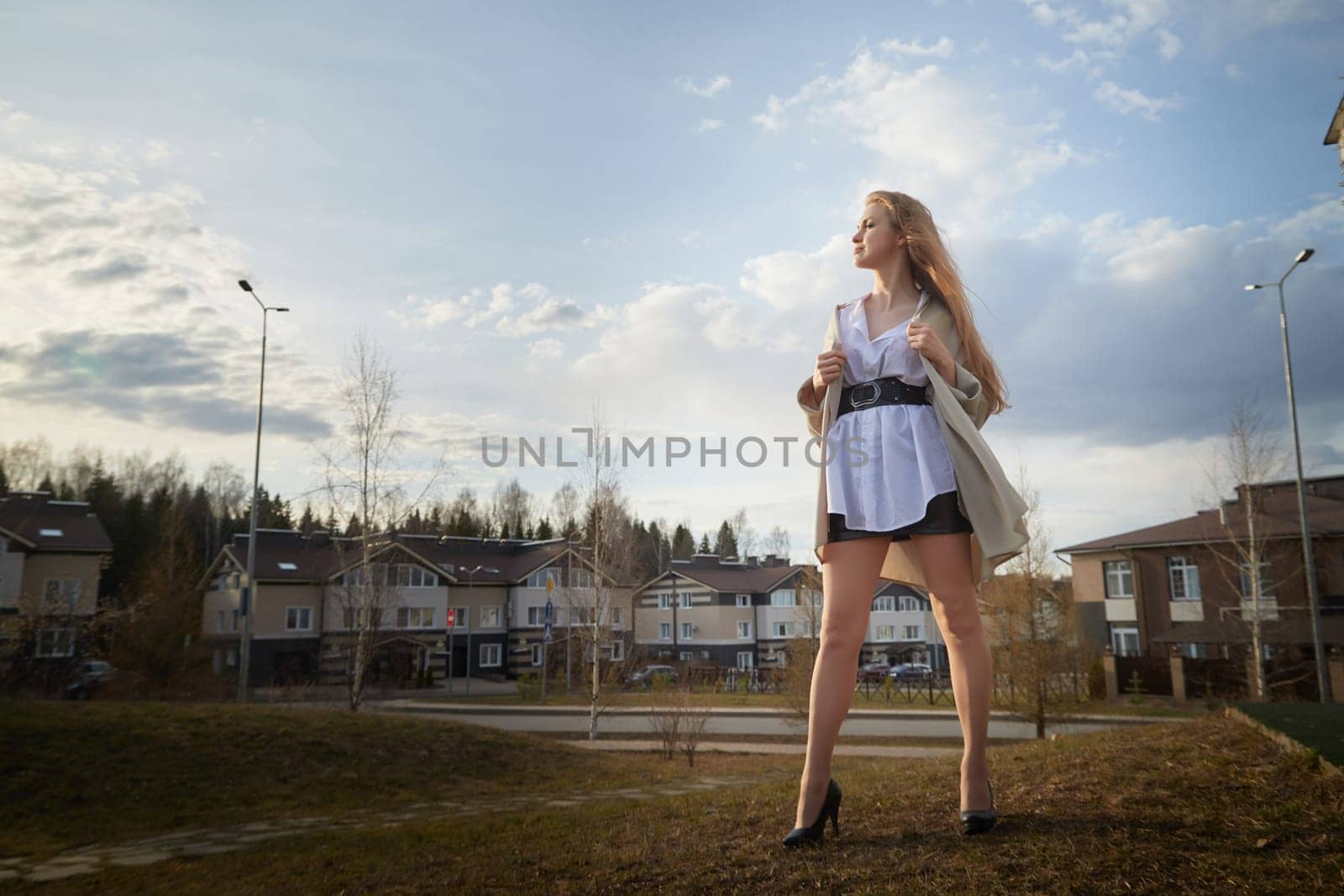 Beautiful girl with long hair in short skirt, white shirt and jacket in village or small town. Tall young slender woman and houses and sky with clouds on background on autumn, spring or summer day