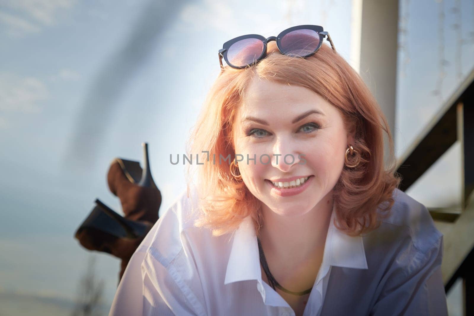 Portrait of Beautiful girl with red hair in white shirt in open wooden pavillion in village or small town. Young slender woman and sky with clouds on background on autumn, spring or summer evening