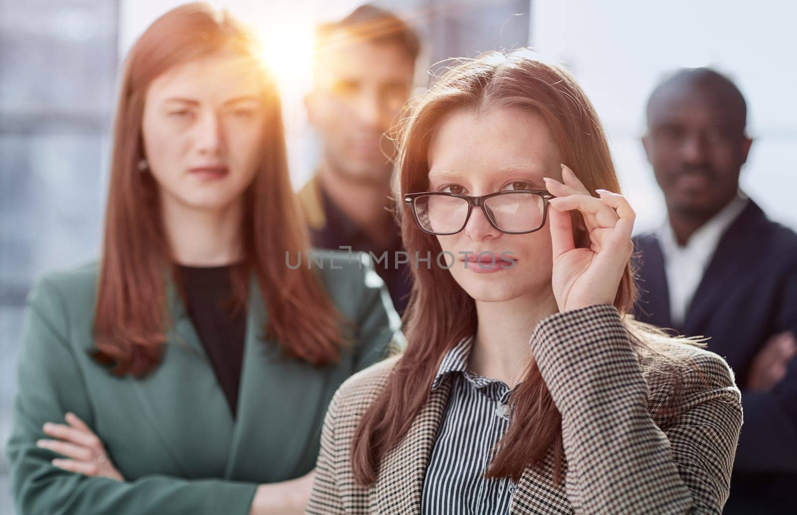 Modern young businesswoman in formal wear adjusting glasses looking at camera while standing in office instead of with colleagues