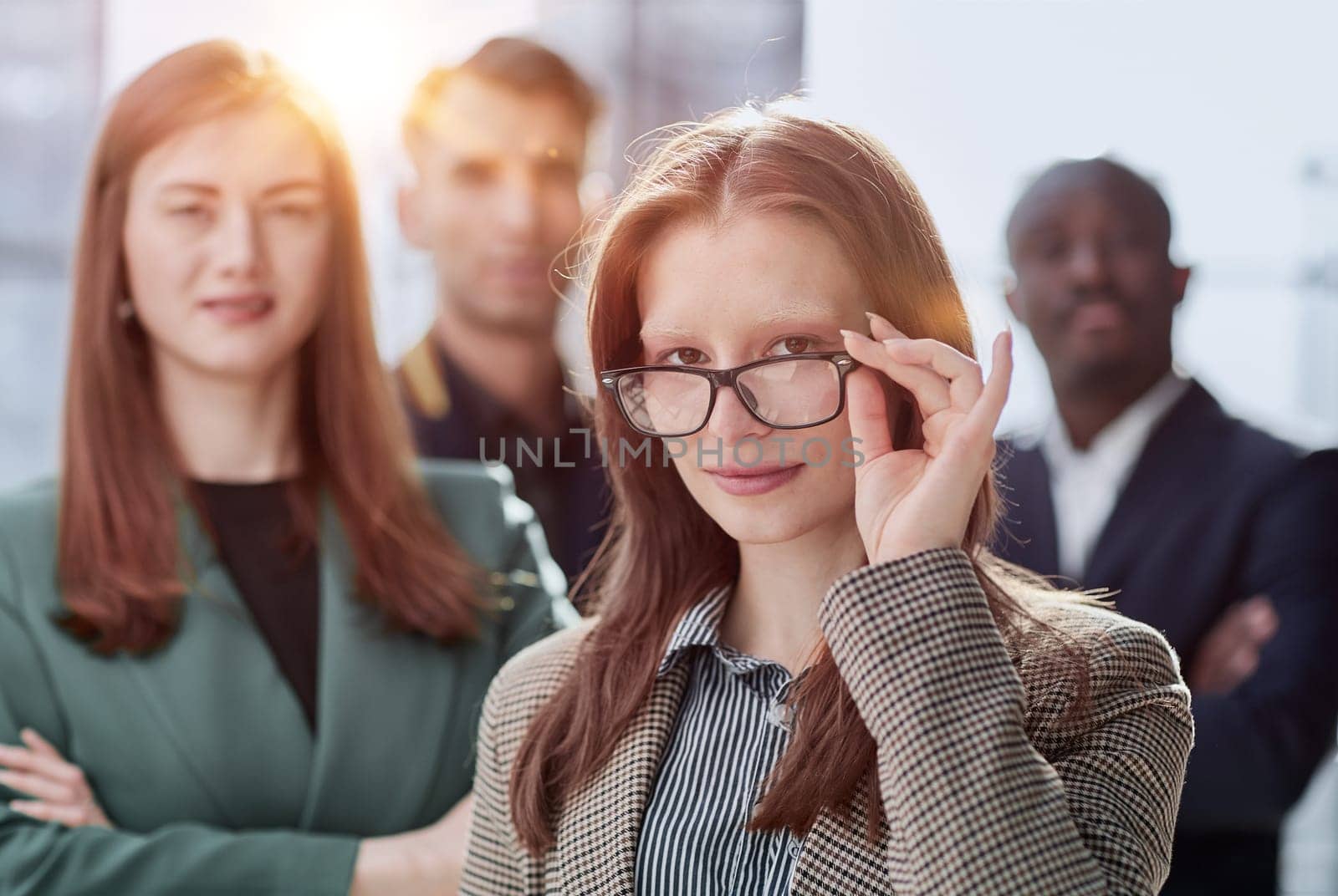 businesswoman in formal wear adjusting her glasses looking at the camera while standing in the office instead of with colleagues by Prosto