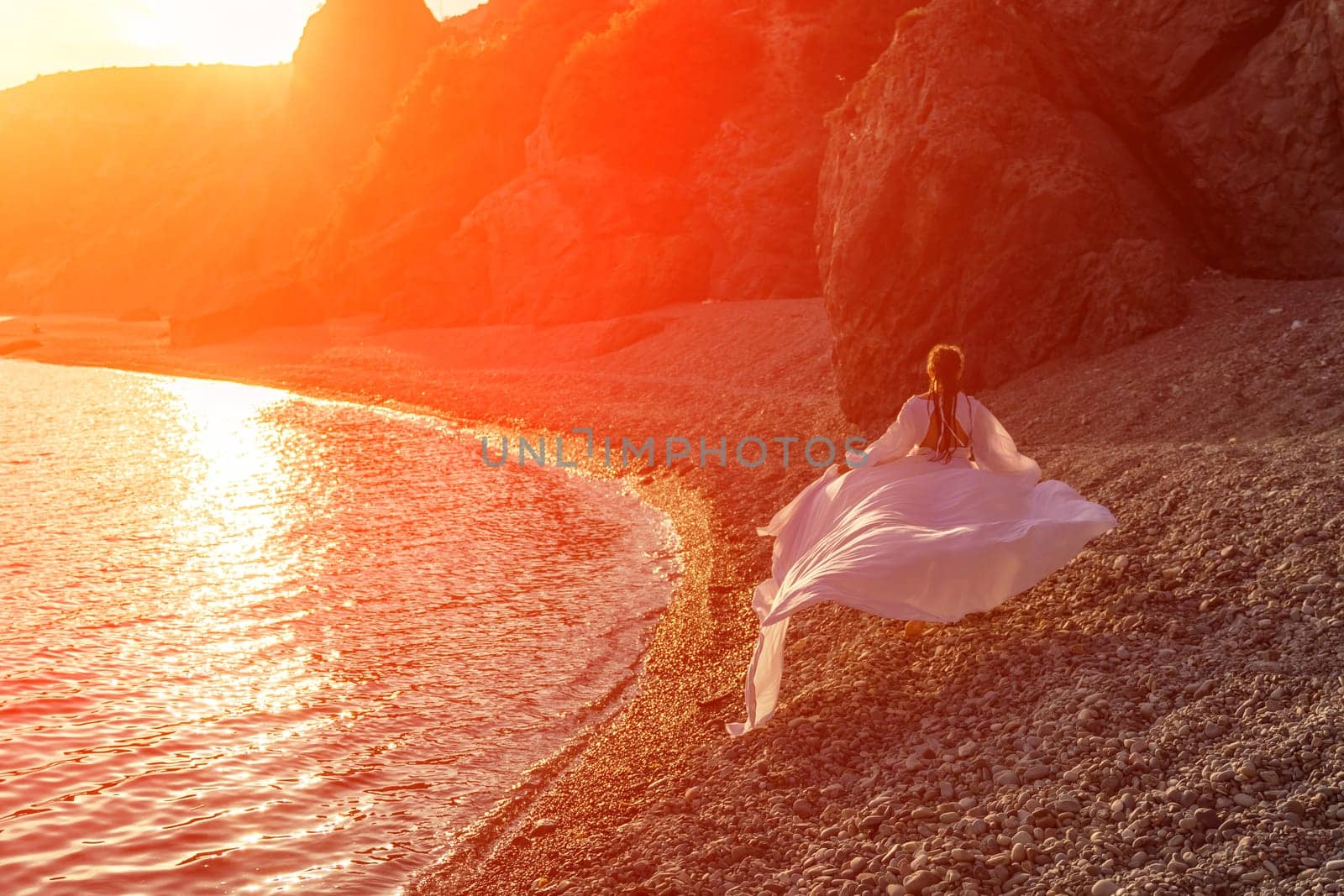 Mysterious woman silhouette long hair walks on the beach ocean water, sea nymph wind listens to the wave. Throws up a long white dress, a divine sunset. Artistic photo from the back without a face.