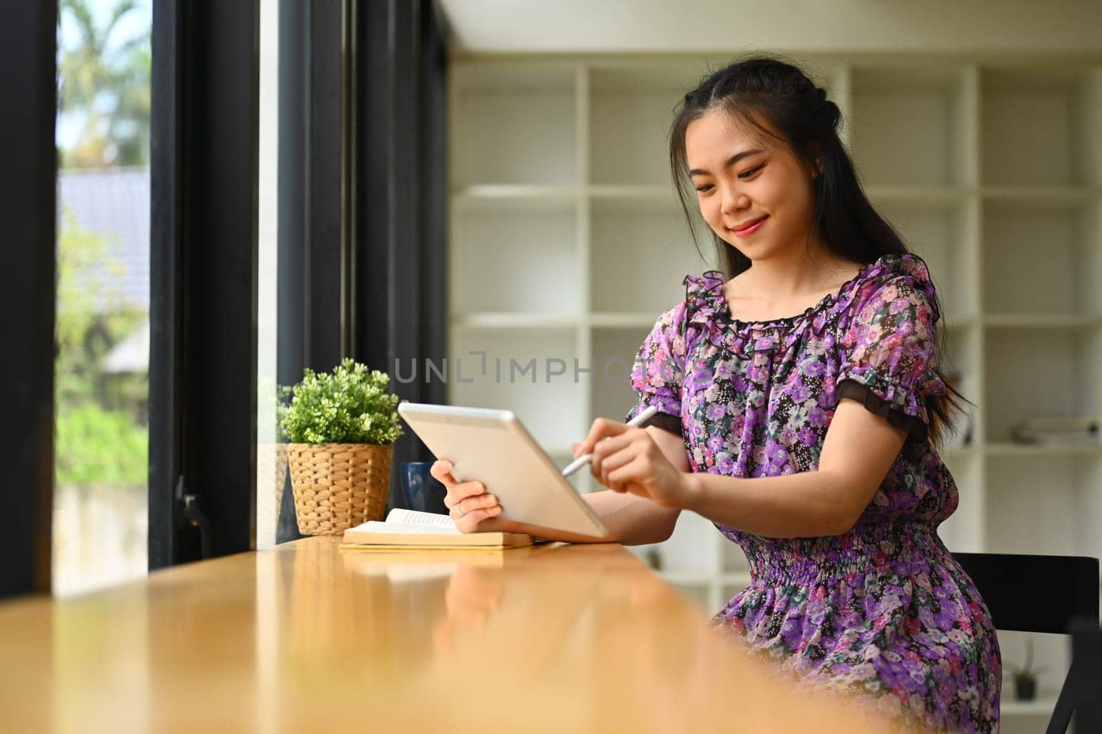 Smiling teenage asian woman using digital tablet on wooden table in coffee shop.