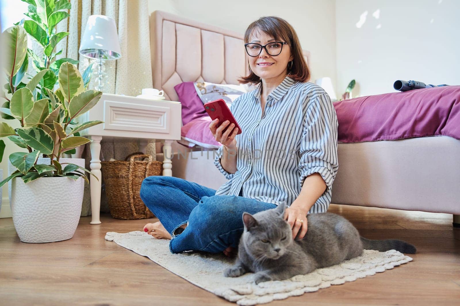 Home lifestyle, woman with cat, comfort calmness concept. Female sitting on floor on carpet using smartphone, pet gray british cat lying near owner