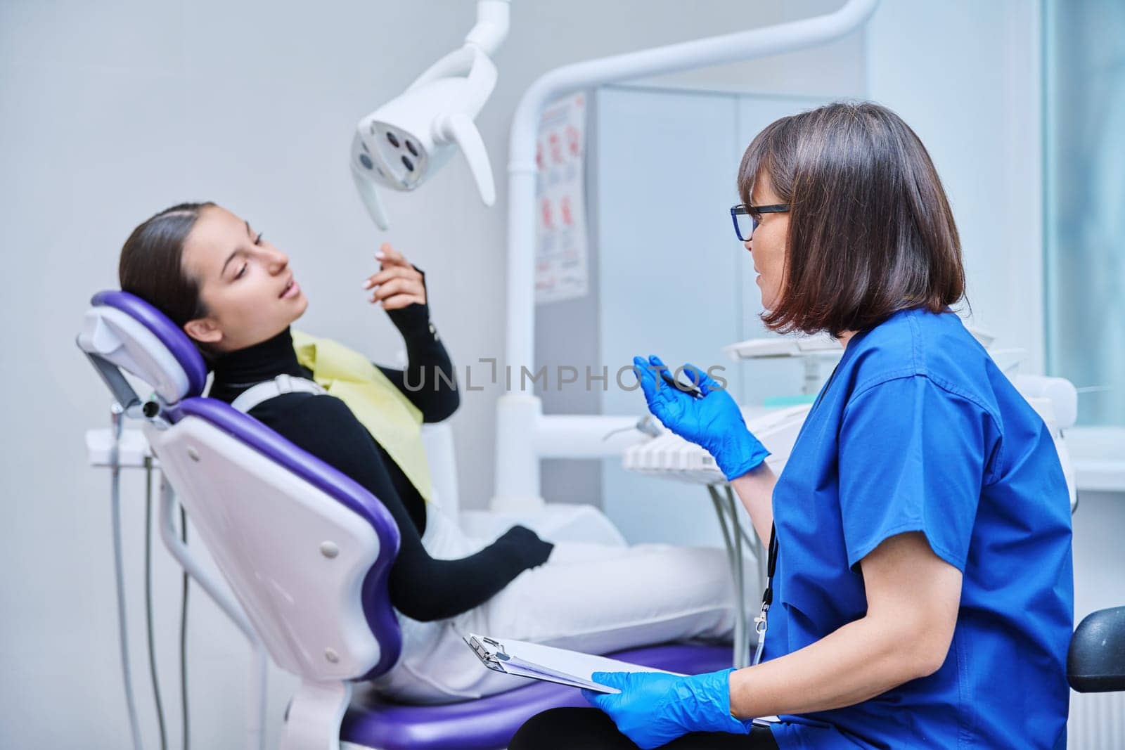 Dentist and teenage girl patient in dental clinic, doctor nurse writing on clipboard consulting, teen female talking about toothache disturbing dental problem. Dentistry, treatment, dental health care