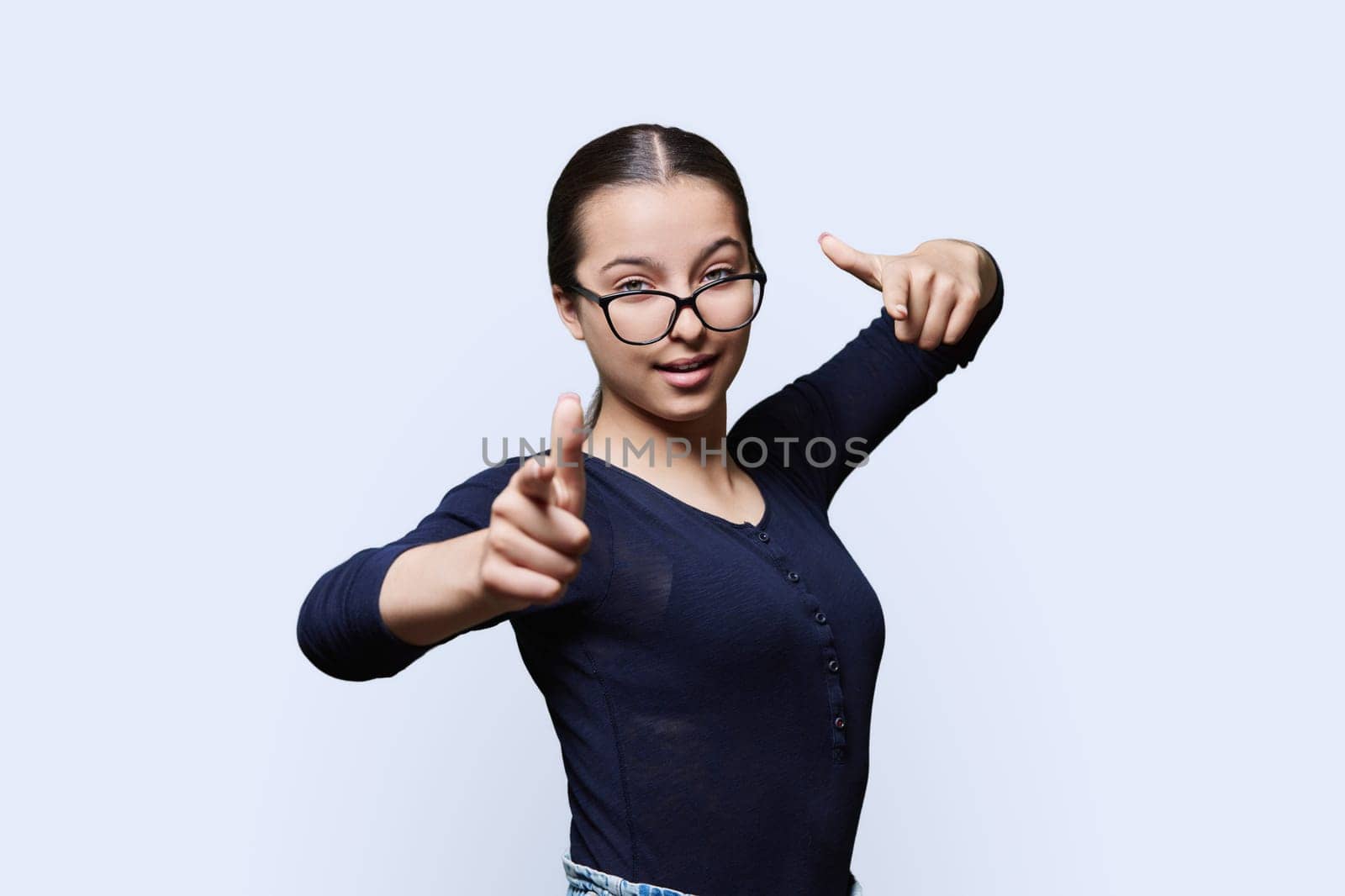 Teenage student girl in glasses looking at camera pointing finger at you, on white studio background. Announcement, highlighting, attention, education, lifestyle people concept