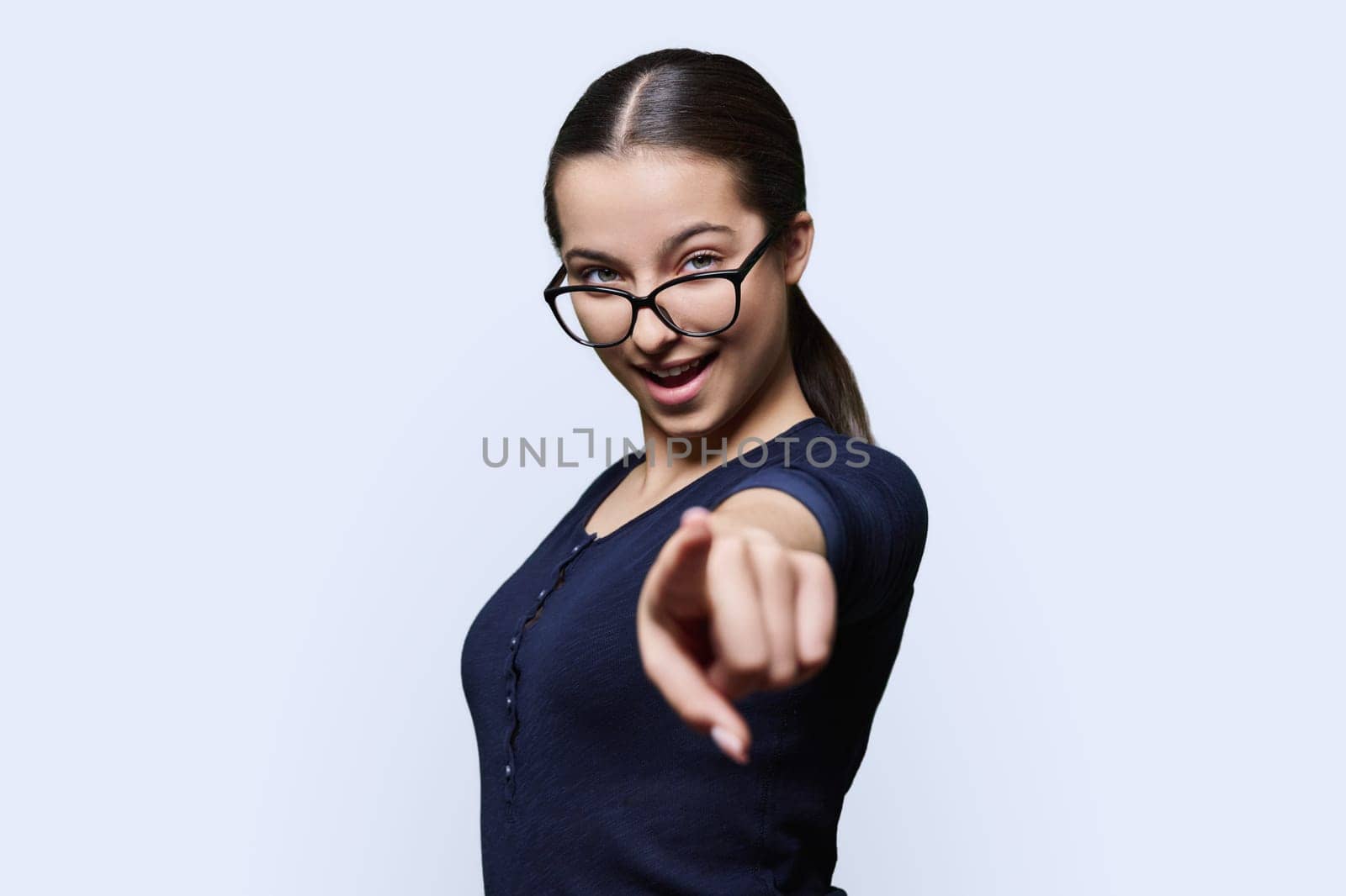 Teenage student girl in glasses looking at camera pointing finger at you, on white studio background. Announcement, highlighting, attention, education, lifestyle people concept