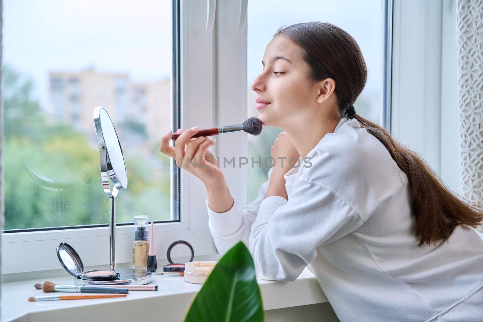 Young teenage girl with cosmetics mirror doing natural makeup, at home near the window, using brushes, cosmetics. Beauty, fashion, youth concept