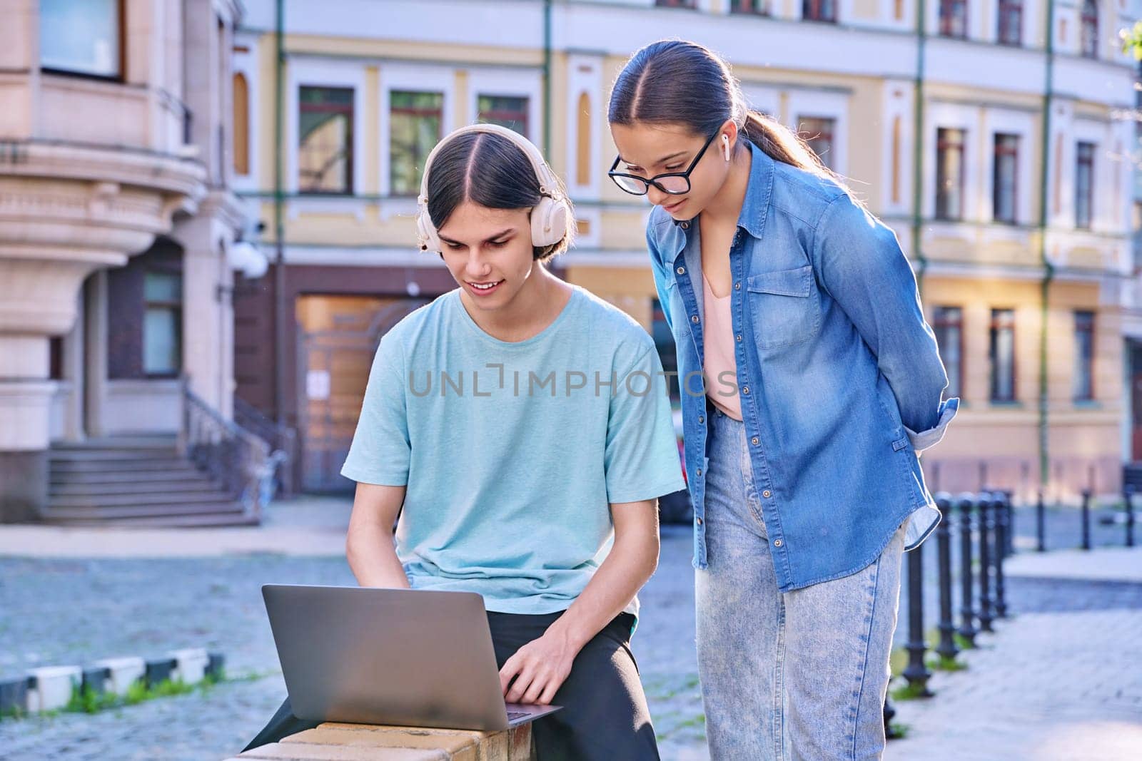 Teenage male and female using laptop for study, leisure, outdoor on city street. Guy and girl teenagers looking together at aptop screen. Lifestyle, technology, youth, education, city life concept