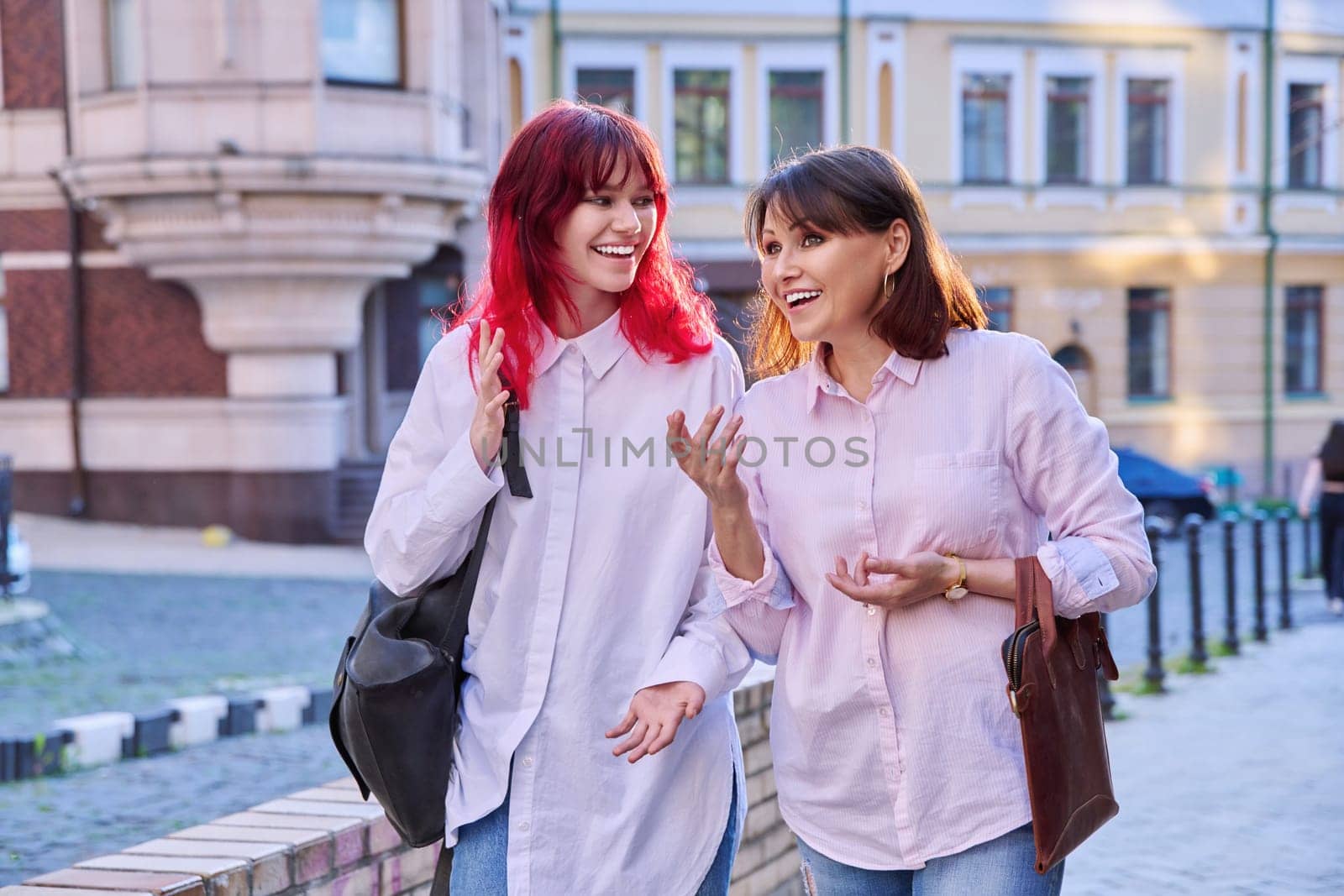 Two generations, smiling joyful happy mother and stylish attractive teenage daughter, walking talking together along city street. Family communication parent child teenager lifestyle leisure people