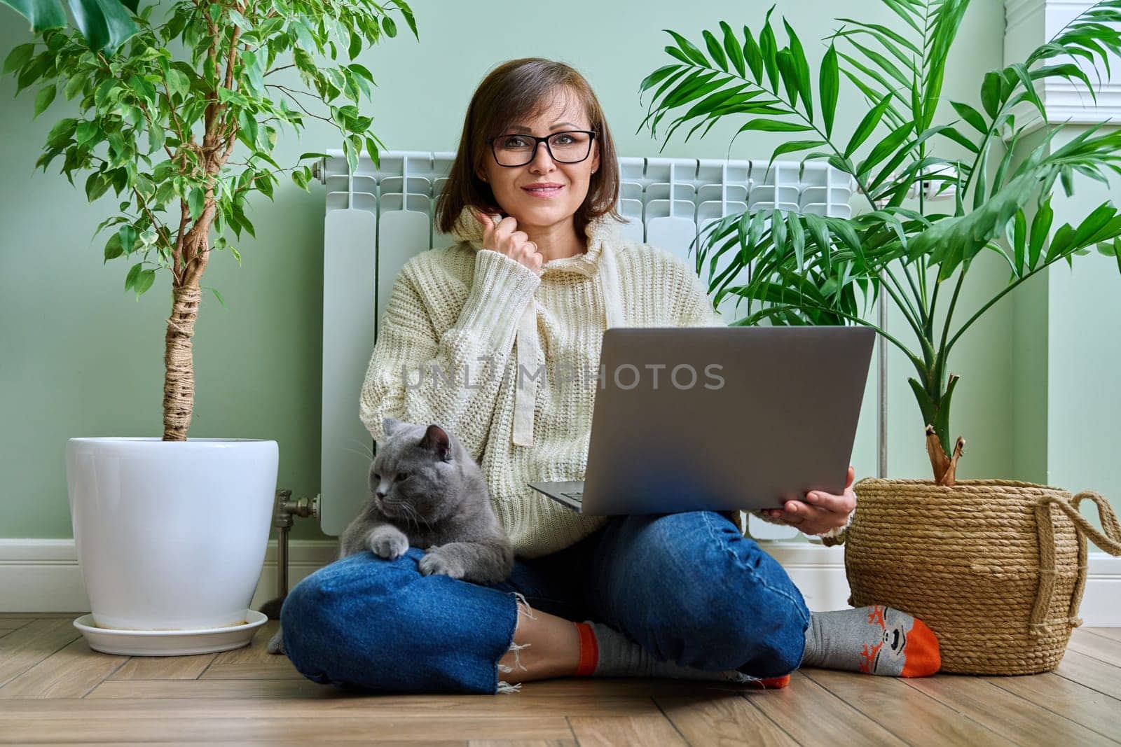 Middle-aged woman in warm clothes with cat using laptop. Female warming at home near heating radiator, with pet in her arms. Cold season winter autumn, leisure lifestyle work freelance, people animals