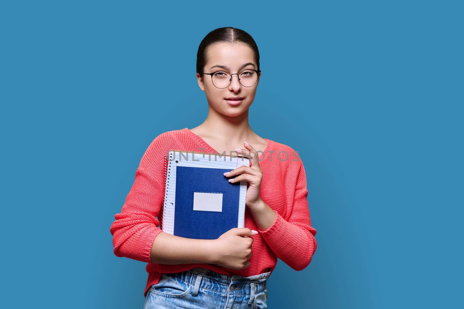 Portrait of teenage female high school student in glasses holding notebooks smiling looking at camera against blue studio background. Education, adolescence, study concept