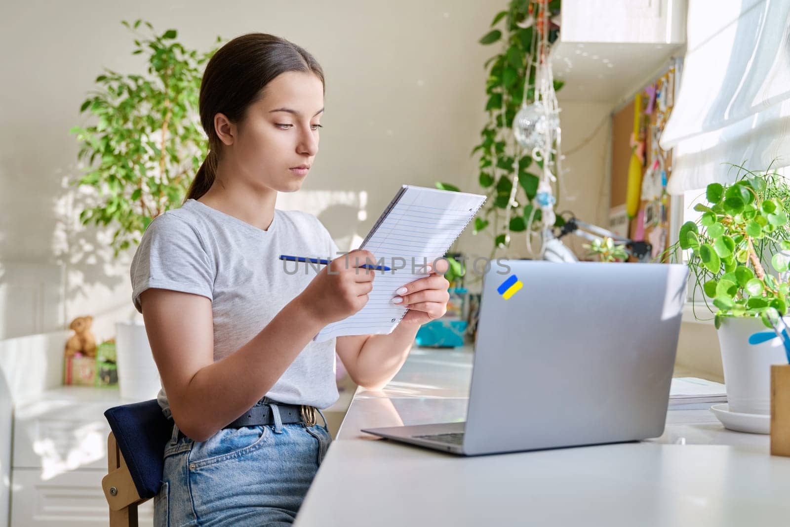 Teenage girl studying at home using laptop making notes in notebook. Ukrainian high school student preparing for control exam, doing homework, writing essay. Education, knowledge, adolescence concept