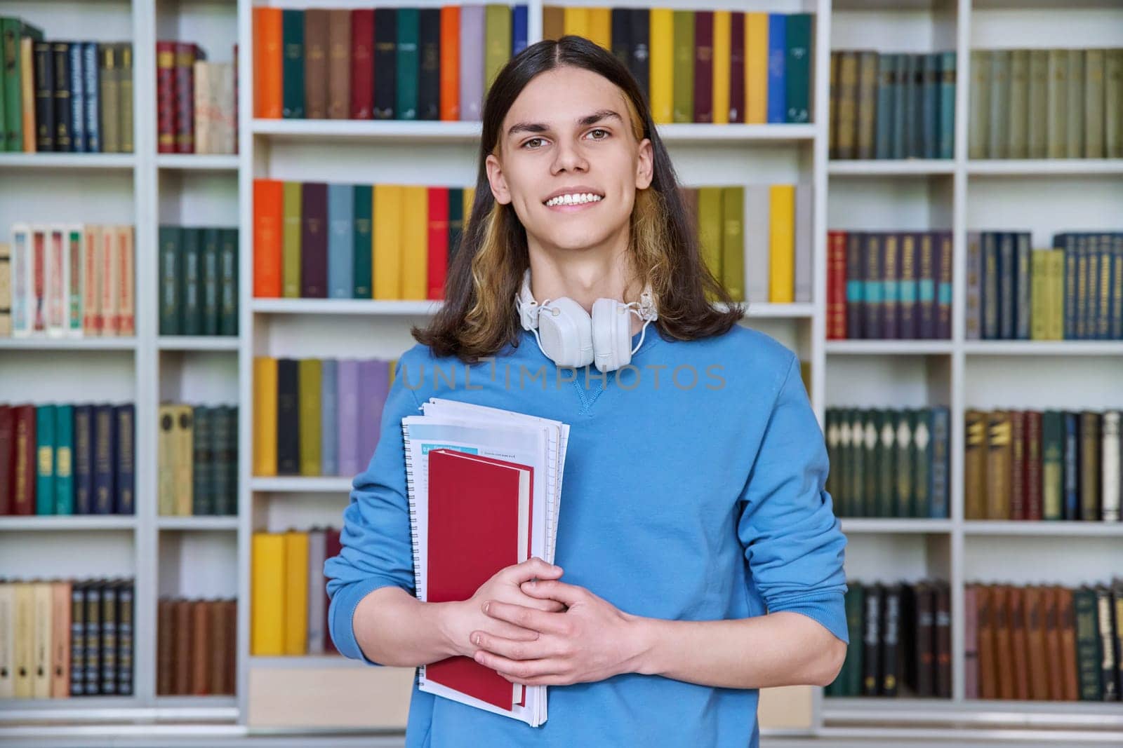 Portrait of teenage student guy looking at camera in library. Smiling male teenager with headphones, textbooks and books in his hands. College, university, education, knowledge, youth concept