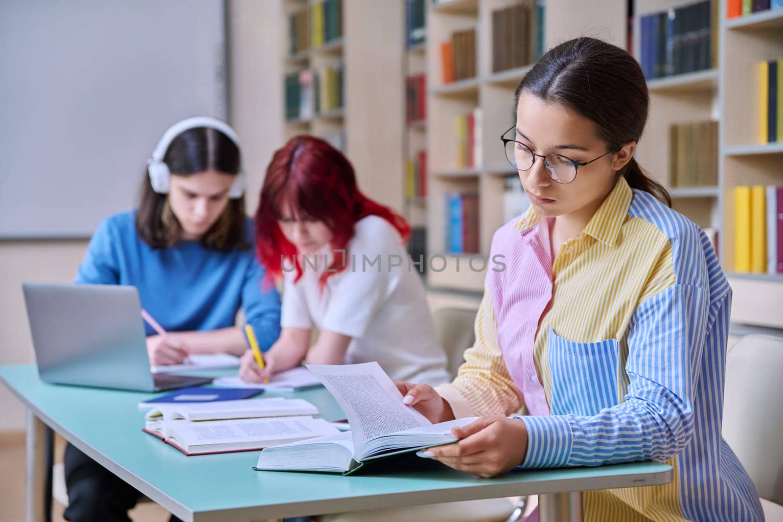 High school students studying in library class, teenage girl in focus. Group of teenagers sitting at desk, writing in notebooks, using laptop books. Knowledge, education, adolescence concept