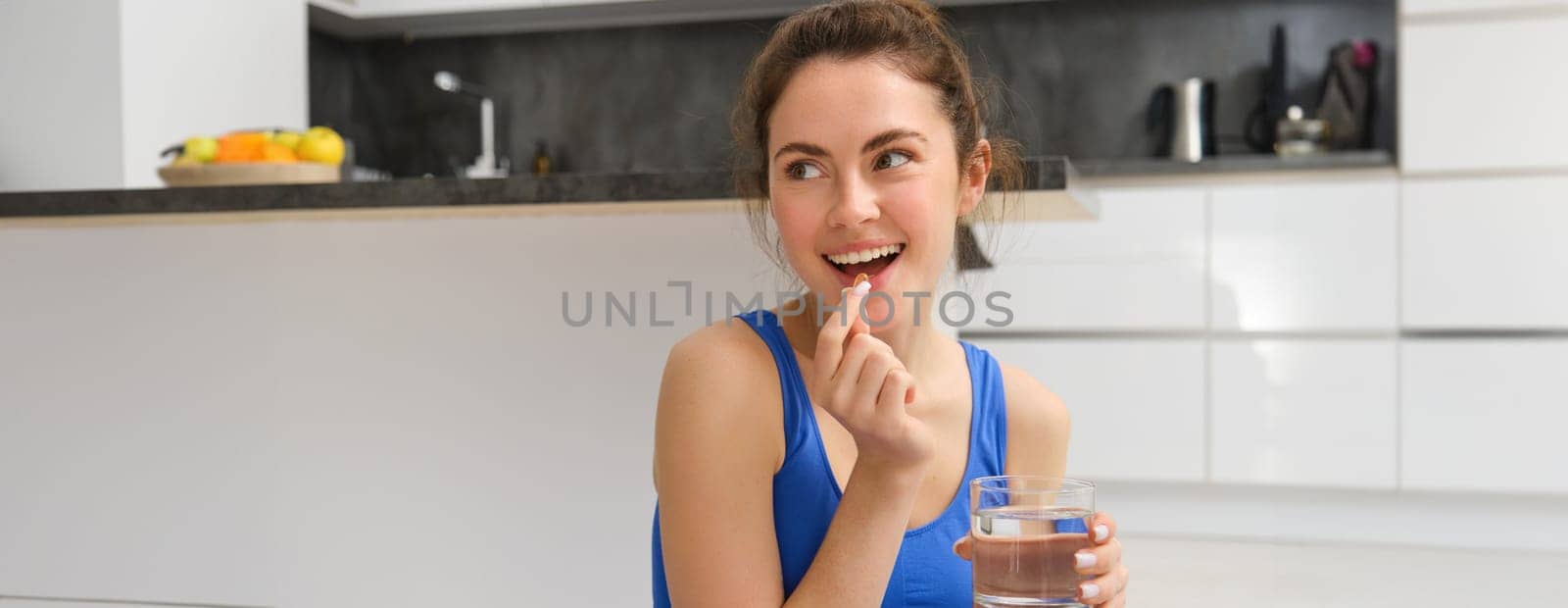 Close up portrait of brunette fitness woman, taking dietary supplements, vitamins and glass of water after workout at home.