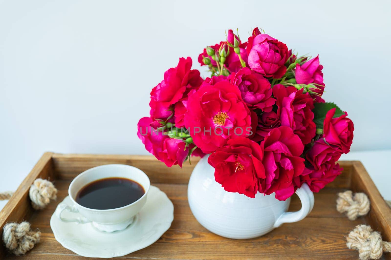 Still life with a cup of coffee on a wooden tray and a vase with freshly cut homemade roses. Romantic breakfast. Eating and drinking in the morning sunlight