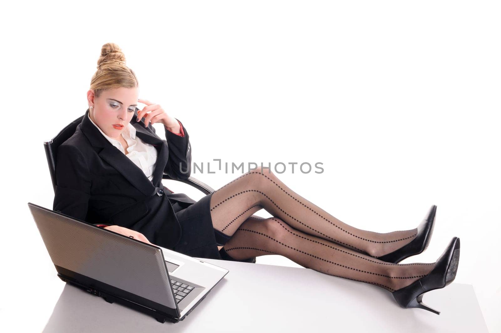 Young woman with blond hair wearing a business suit and has bored in the office. You sitting bored on the laptop and has her feet on the desk, isolated against white background.