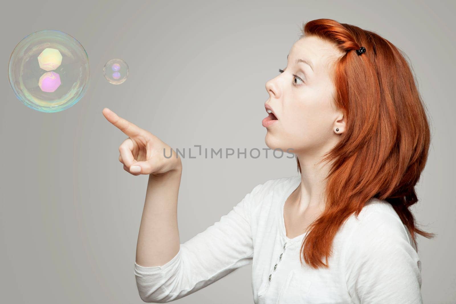 young beautiful red haired girl look to soap bubbles and wondering in front of grey background