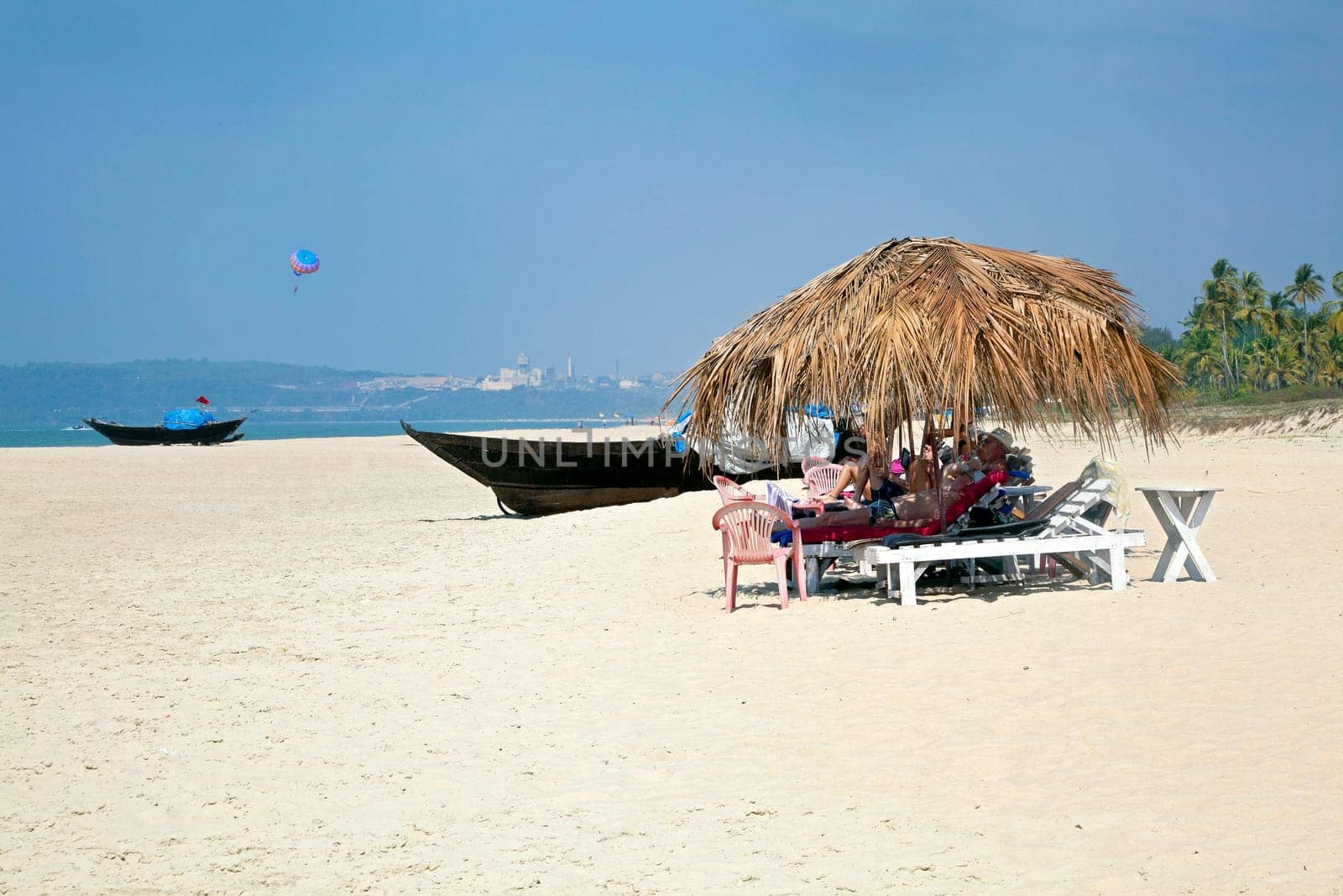 Landscape of secluded beach with day trippers and holidaymakers basking in the sun under the shade of coconut parasols. White sand, blue sky, sea, surf and fishing boats for a backdrop. Generic tropical shot location Uttorda Beach, Goa India