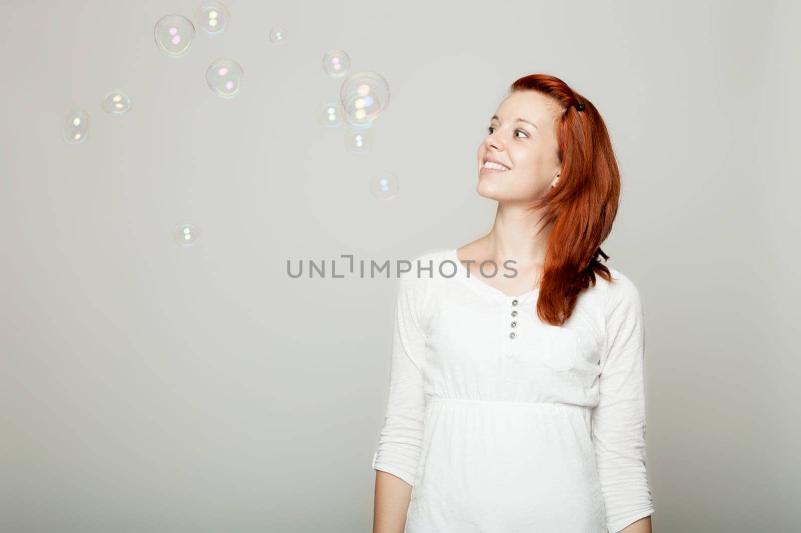 young beautiful red haired girl look to soap bubbles and dream in front of grey background