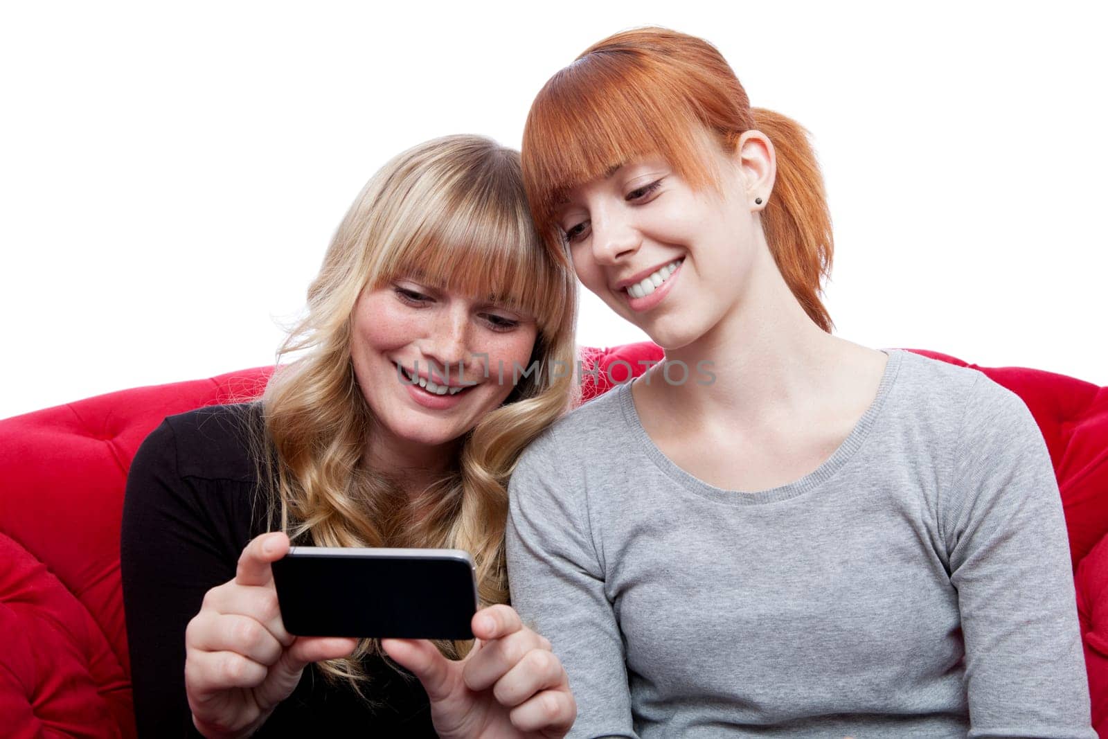 young beautiful blond and red haired girls showing something on cell phone on red sofa in front of white background