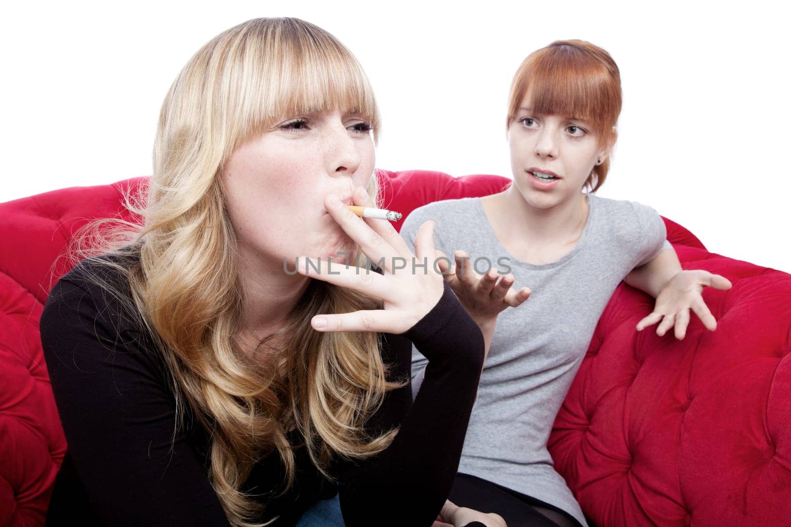 young beautiful blond and red haired girls is shocked about friend smoking on red sofa in front of white background