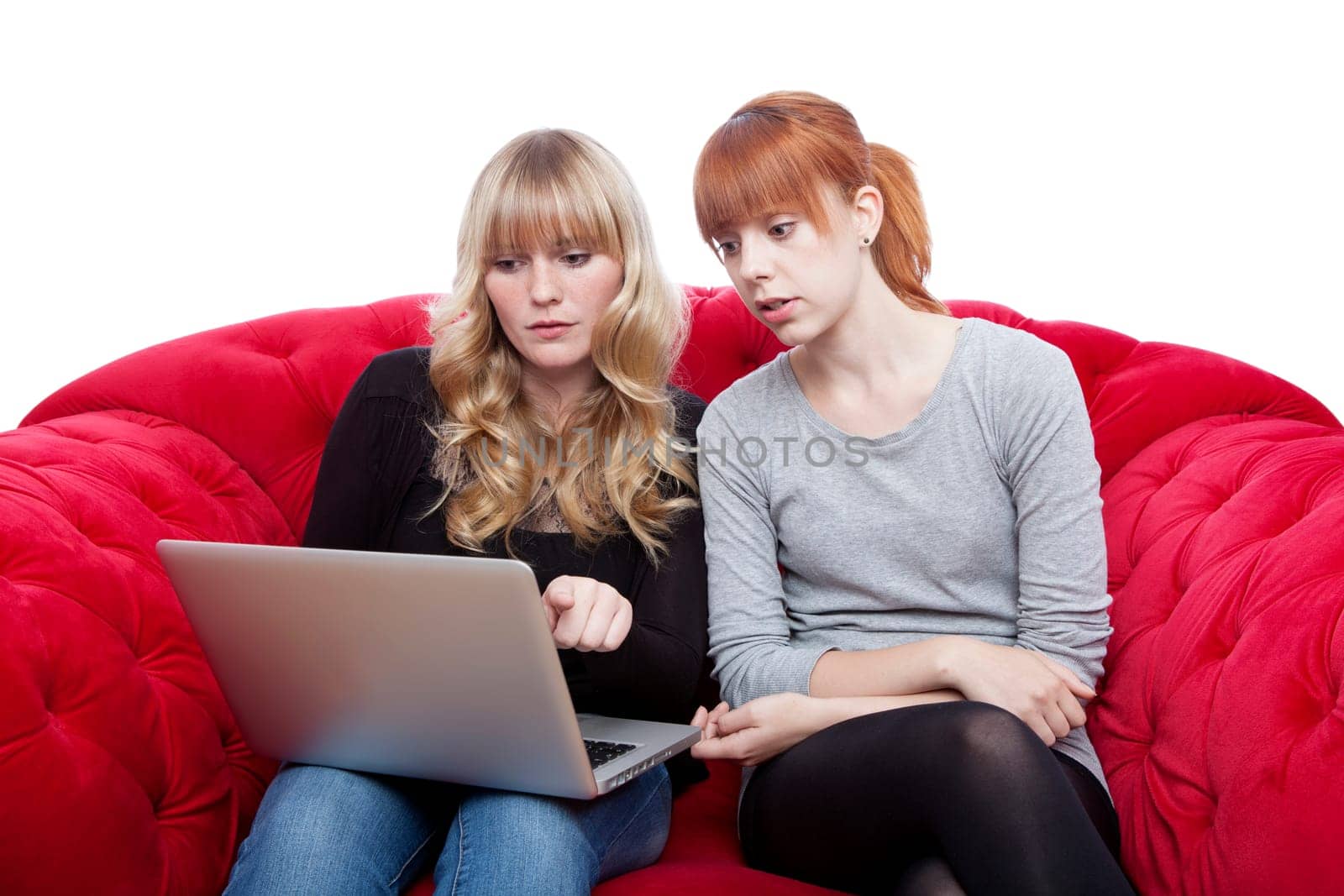 young beautiful blond and red haired girls on red sofashowing something on laptop in front of white background