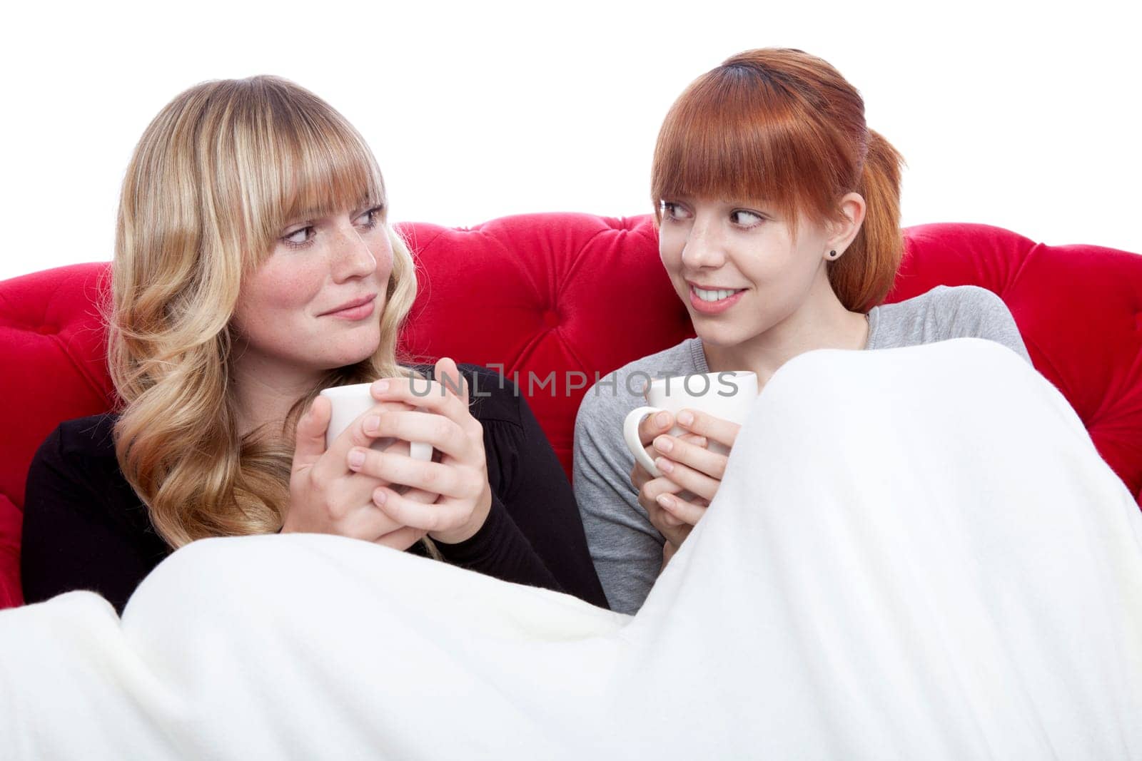 young beautiful blond and red haired girls on red sofa with coffee under blanket in front of white background