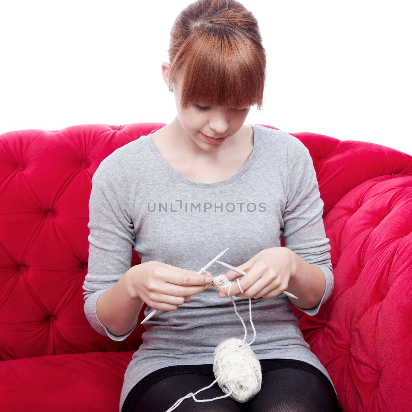 young beautiful red haired girl knitting on red sofa in front of white background