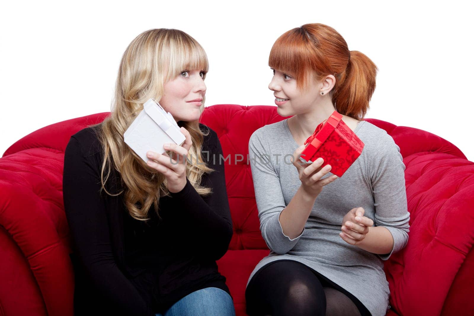 young beautiful blond and red haired girls are happy about gifts on red sofa in front of white background
