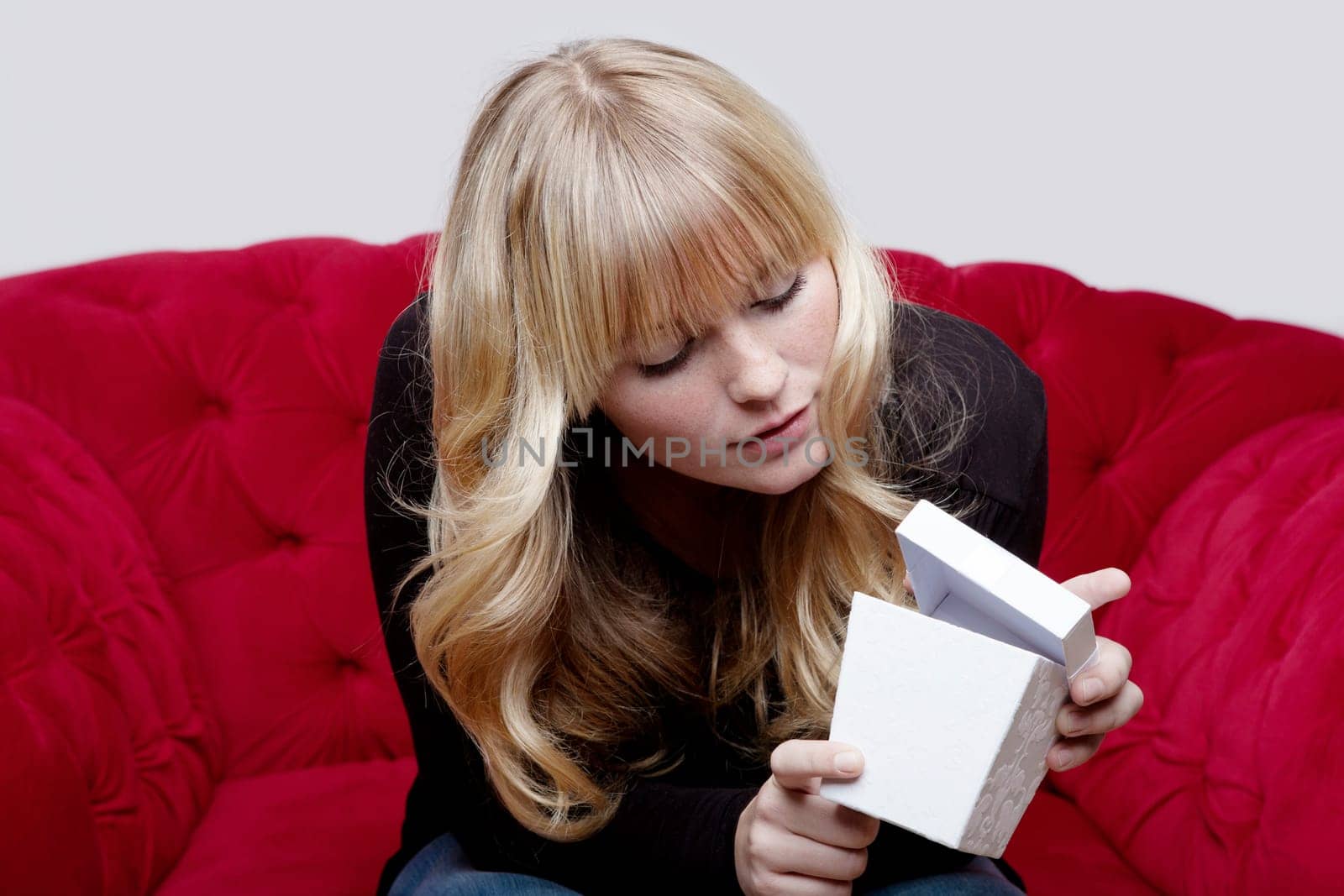 young blond haired girl with present box on red sofa in front of grey background