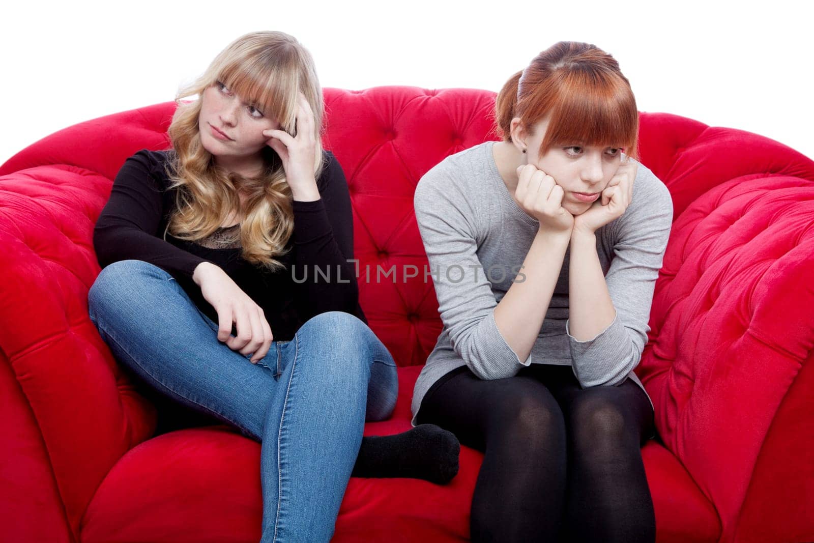 young beautiful blond and red haired girls are bored and depressed on red sofa in front of white background